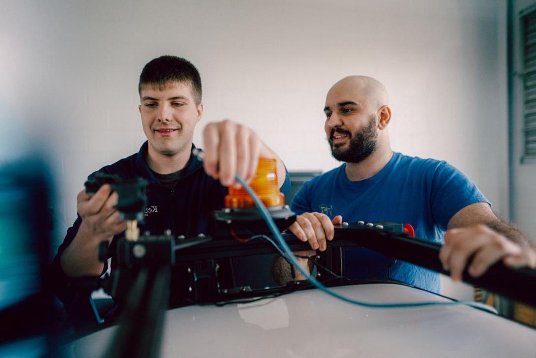 Two Kettering students stand behind a metal frame. One holds a blue wire and prepares to insert it into a mechanical part.