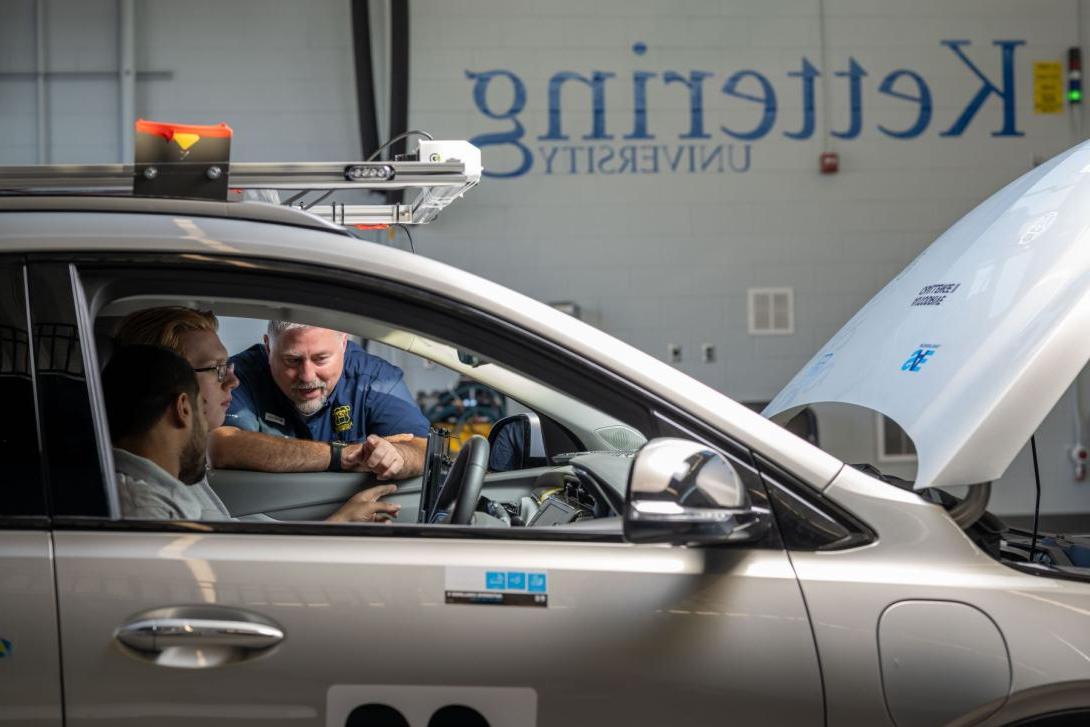 Two Kettering students and a professor look at a car's computer panel in the Mobility Research Center. The car's hood is up